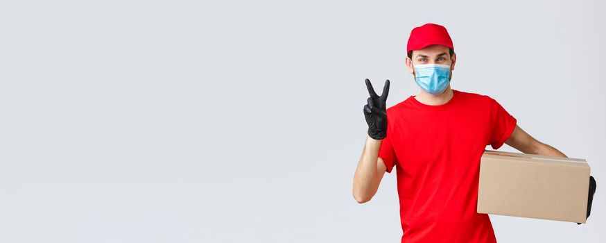 Packages and parcels delivery, covid-19 quarantine delivery, transfer orders. Friendly courier in red uniform, face mask and gloves, deliver order to client, holding box, show peace sign.