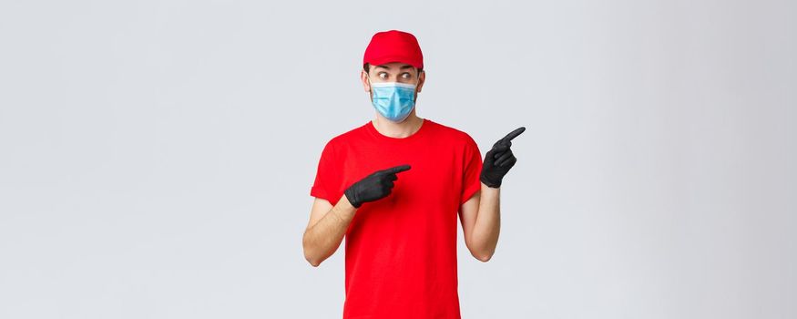 Covid-19, self-quarantine, online shopping and shipping concept. Shocked and impressed delivery guy in medical mask and gloves, pointing right and staring at promo speechless, wear uniform.