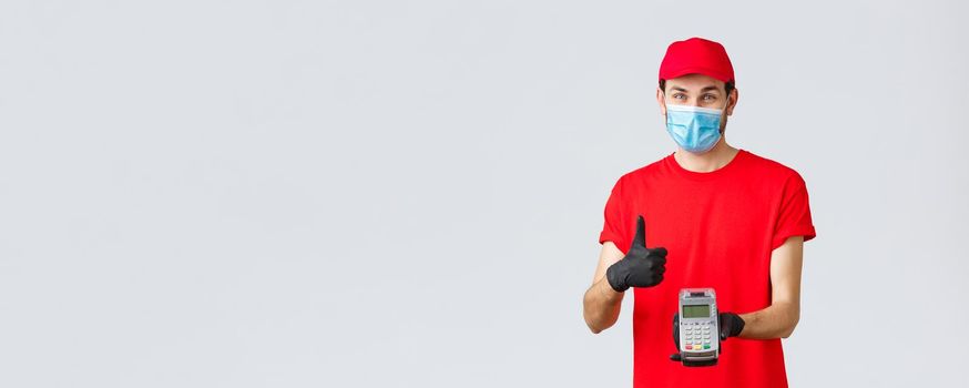 Contactless delivery, payment and online shopping during covid-19, self-quarantine. Friendly smiling courier in red uniform cap, t-shrit, medical mask and gloves, advice pay order with POS terminal.