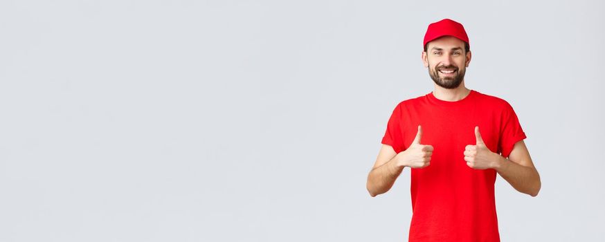 Online shopping, delivery during quarantine and takeaway concept. Cheerful courier in red uniform cap and t-shirt, recommends make orders, thumbs-up in approval, grey background.