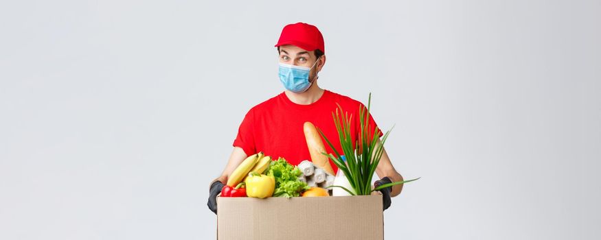 Groceries and packages delivery, covid-19, quarantine and shopping concept. Friendly courier in face mask and gloves, red uniform bring food box to customer ordered online, contactless deliver.