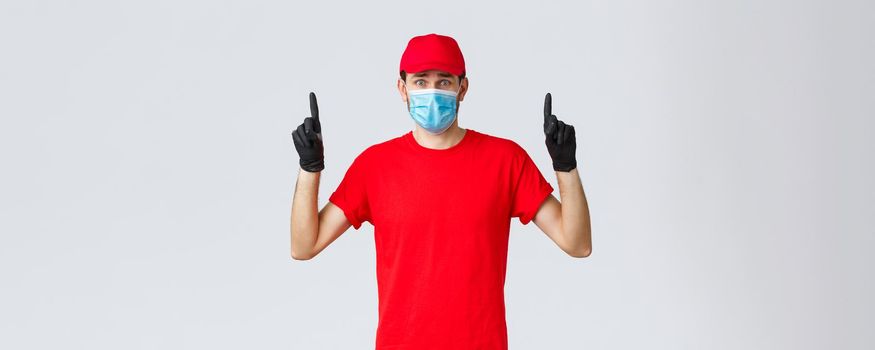 Covid-19, self-quarantine, online shopping and shipping concept. Nervous or scared, worried delivery man in red uniform cap, t-shirt, frowning pointing fingers up at bad news, wear mask and gloves.