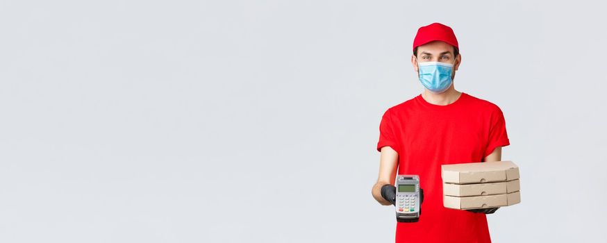 Food delivery, application, online grocery, contactless shopping and covid-19 concept. Friendly courier in red uniform, face mask and gloves, holding order pizza boxes and give client POS terminal.