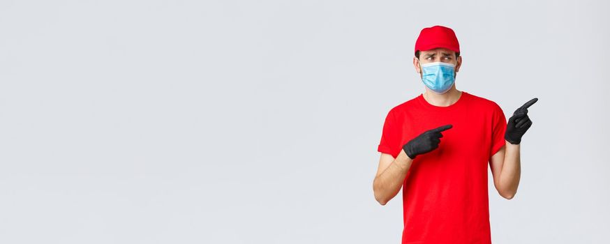 Covid-19, self-quarantine, online shopping and shipping concept. Upset and displeased delivery guy staring and pointing right with reluctant bothered face, wear uniform, medical mask and gloves.