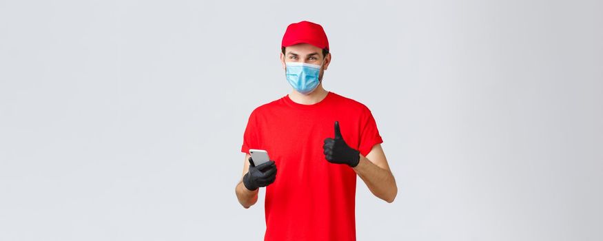 Contactless delivery, payment and online shopping during covid-19, self-quarantine. Friendly courier in gloves and red uniform cap, face mask, show thumb-up as recommend smartphone app or site.