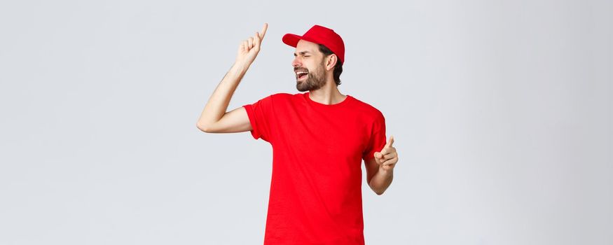 Carefree handsome bearded guy, delivery employee in red t-shirt and cap, singing with closed eyes, raising finger up as reaching high pitch note, standing upbeat over grey background.