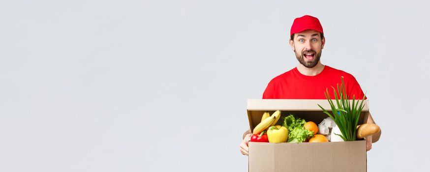 Groceries and packages delivery, covid-19, quarantine and shopping concept. Smiling handsome bearded courier in red uniform, bring food package, grocery order to client in box, look amused.