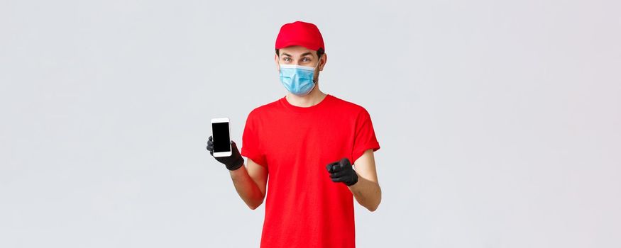 Contactless delivery, payment and online shopping during covid-19, self-quarantine. Smiling courier in red uniform, gloves and face mask, showing order info on phone screen, pointing camera.