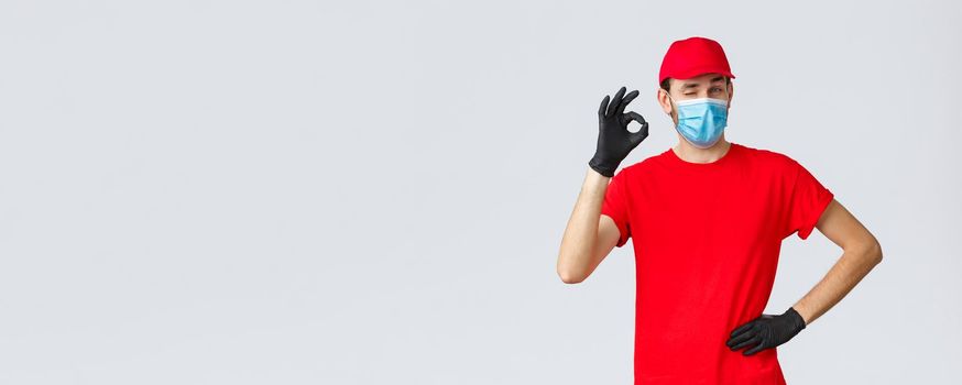 Covid-19, self-quarantine, online shopping and shipping concept. Delivery guy in red t-shirt, cap with face mask and gloves, show okay sign, wink, say no problem. Courier deal with any transfer.