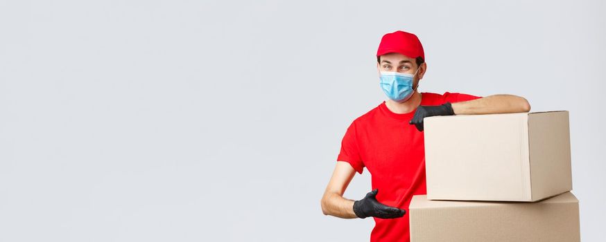 Packages and parcels delivery, covid-19 quarantine and transfer orders. Smiling courier in red uniform, gloves and medical face mask, introduce boxes to transfer your order, recommend service.