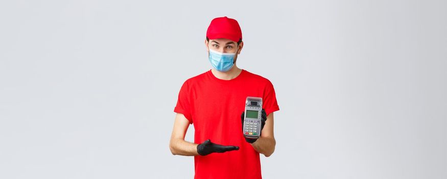 Contactless delivery, payment and online shopping during covid-19, self-quarantine. Pleasant courier in red uniform, face mask and gloves, asking use POS terminal to pay order.