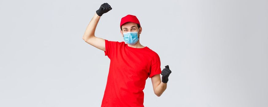 Groceries and packages delivery, covid-19, quarantine and shopping concept. Cheerful courier red uniform, gloves and face mask, raising hands up, chanting rejoicing over good news, fight coronavirus.