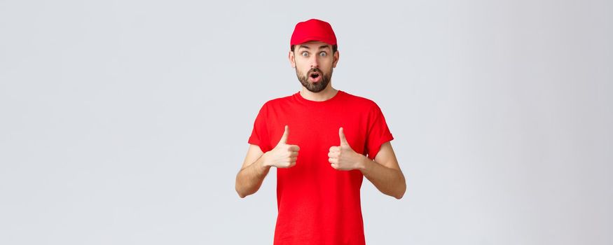 Online shopping, delivery during quarantine and takeaway concept. Impressed and shocked bearded courier in red t-shirt and cap uniform, gasping open mouth astonished, thumbs-up, recommend service.
