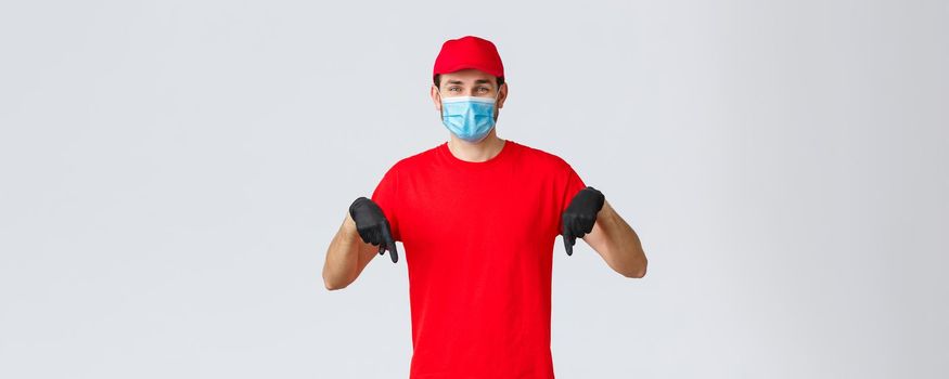 Covid-19, self-quarantine, online shopping and shipping concept. Smiling friendly delivery guy red uniform, cap and t-shirt, wear medical mask with gloves, pointing down, showing promo or client info.