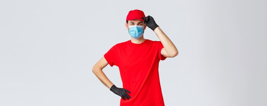 Covid-19, self-quarantine, online shopping and shipping concept. Delivery guy red uniform, touching cap as saluting customer, working in coronavirus outbreak, wear medical mask and protective gloves.