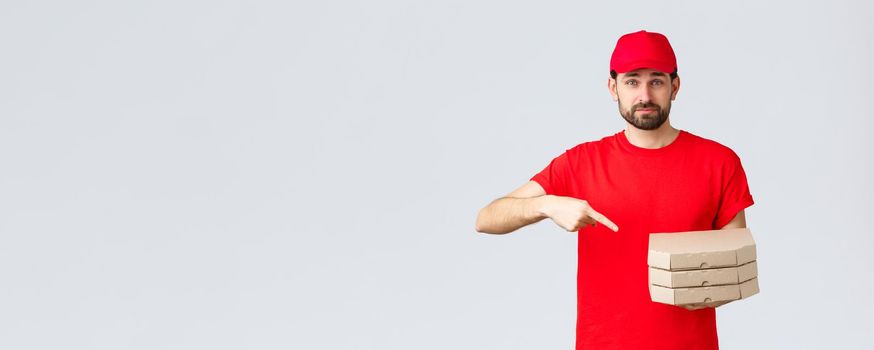 Food delivery, quarantine, stay home and order online concept. Confident friendly courier in red uniform cap and t-shirt, employee bring order pizza, pointing finger at boxes, grey background.