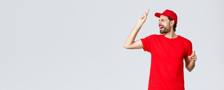 Carefree handsome bearded guy, delivery employee in red t-shirt and cap, singing with closed eyes, raising finger up as reaching high pitch note, standing upbeat over grey background.