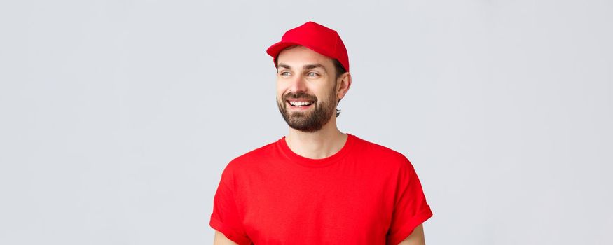 Online shopping, delivery during quarantine and takeaway concept. Cheerful bearded guy in red uniform cap and t-shirt, looking away with pleased smile, reading banner sign, grey background.