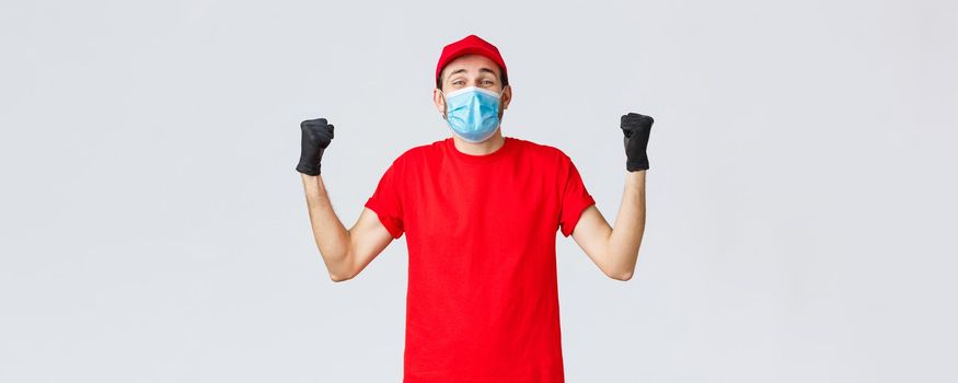 Covid-19, self-quarantine, online shopping and shipping concept. Excited delivery man feel rejoice and happiness, fist pump in celebration, success, achieve goal, wear medical mask and uniform cap.