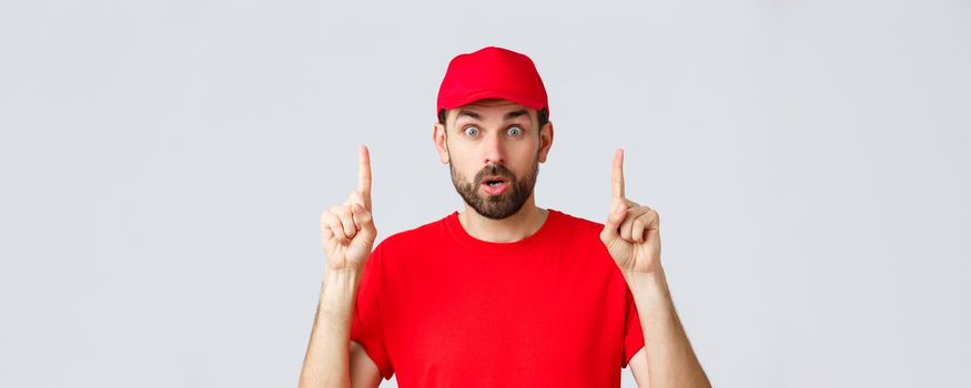 Online shopping, delivery during quarantine and takeaway concept. Surprised and impressed delivery guy, courier in red t-shirt and cap, pointing fingers up, gasping amazed, read interesting info.