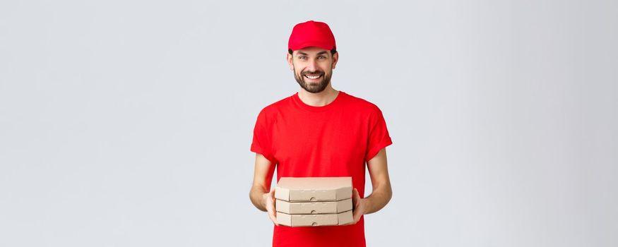 Food delivery, quarantine, stay home and order online concept. Friendly smiling courier in red uniform giving client order. Employee bring pizza at your house, standing grey background.