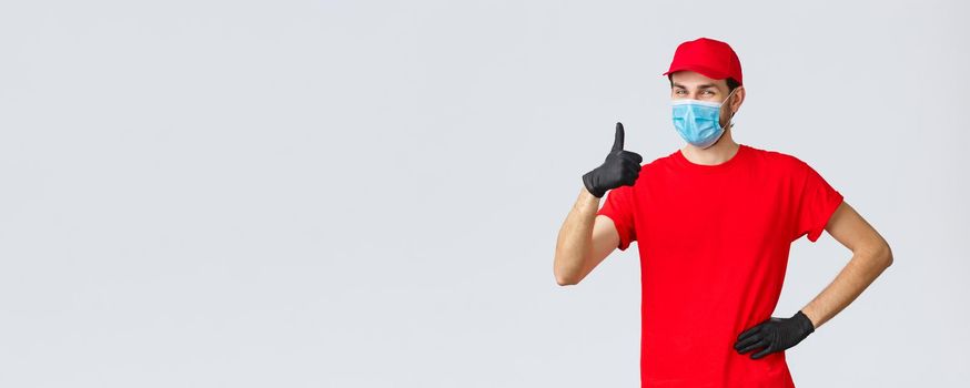 Covid-19, self-quarantine, online shopping and shipping concept. Friendly cheerful delivery guy in red uniform, gloves and face mask guarantee courier service quality, show thumb-up in like, approval.