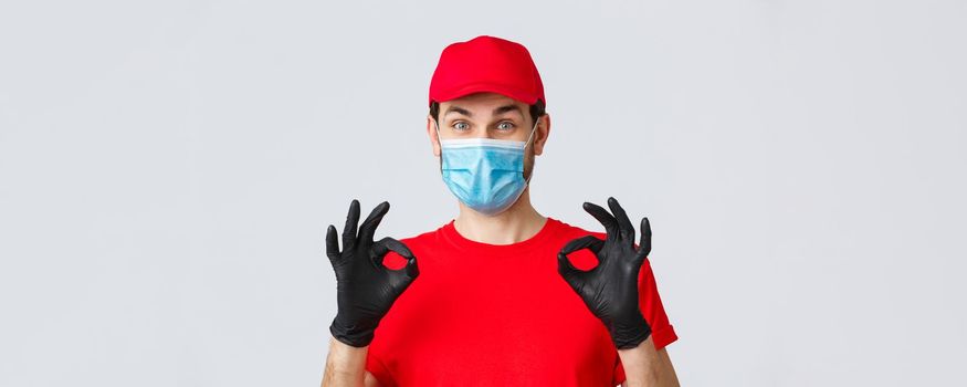 Contactless delivering, covid-19 and shopping concept. Cheerful, pleased courier in red uniform, cap and medical mask with gloves show okay, approval or guarantee gesture, recommend service.