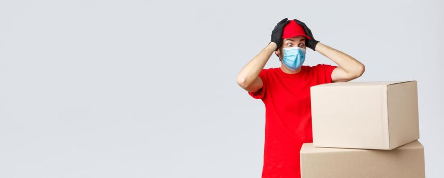 Packages and parcels delivery, covid-19 quarantine and transfer orders. Concerned and troubled courier in red uniform, face mask and gloves, grab head and gasping shocked staring at boxes.