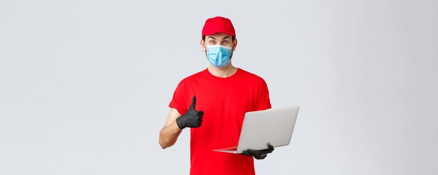 Customer support, covid-19 delivery packages, online orders processing concept. Enthusiastic courier in red uniform thumbs-up reading promo on webpage. Delivery guy hold laptop, wear mask and gloves.