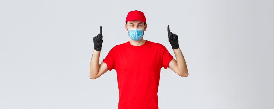 Covid-19, self-quarantine, online shopping and shipping concept. Surprised delivery guy in red uniform and cap, raising eyebrows amazed or excited, showing promo top, pointing up, wear medical mask.