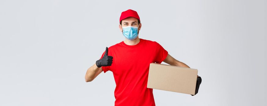 Packages and parcels delivery, covid-19 quarantine delivery, transfer orders. Cheerful courier in red uniform, gloves and face mask, thumb-up, recommend contactless deliver, holding box with order.