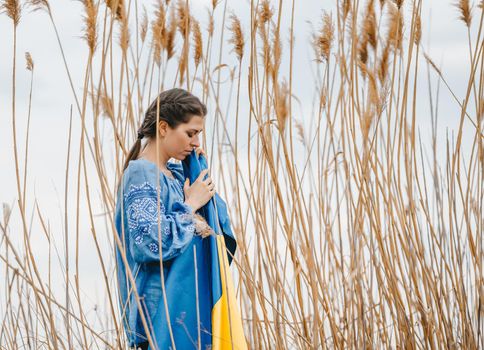 Sorrowful ukrainian woman with national flag on natural reeds background. Lady in blue embroidery vyshyvanka blouse. Ukraine, independence, freedom, patriot symbol, victory in war. High quality photo