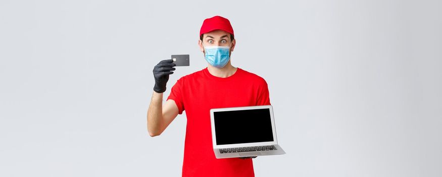 Customer support, covid-19 delivery packages, online orders processing concept. Surprised excited courier in red uniform, face mask and gloves, showing credit card to pay orders and laptop.