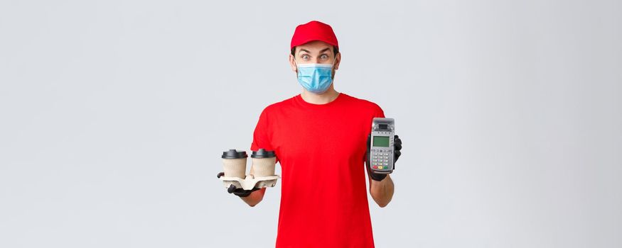 Takeaway, food and groceries delivery, covid-19 contactless orders concept. Excited and surprised client or employee in red uniform, face mask and gloves, giving client paying terminal with coffee.