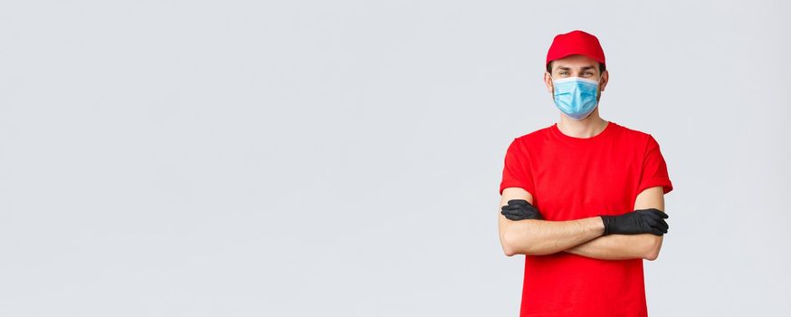 Covid-19, self-quarantine, online shopping and shipping concept. Confident smiling delivery man in red cap, t-shirt, wearing protective medical mask and rubber gloves while making courier order.