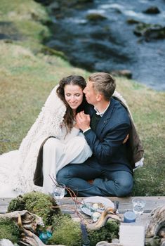 Groom kisses bride in a blanket at the laid table on the river bank. High quality photo