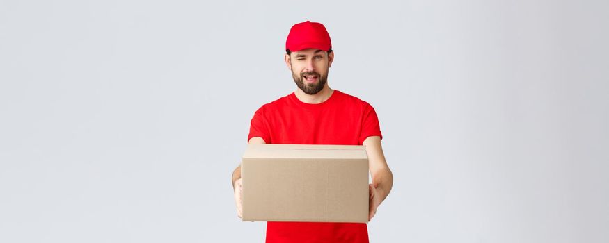 Order delivery, online shopping and package shipping concept. Cheeky handsome bearded courier in red uniform, handing box package to client. Employee wink to you and give order parcel.