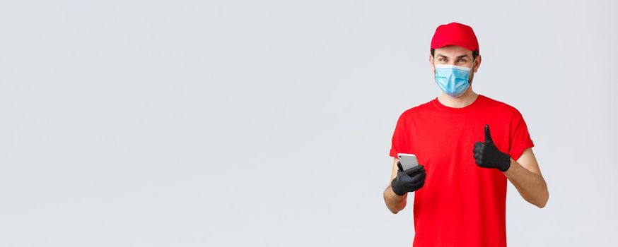 Contactless delivery, payment and online shopping during covid-19, self-quarantine. Friendly courier in gloves and red uniform cap, face mask, show thumb-up as recommend smartphone app or site.