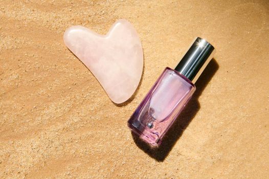 Flat lay. Minimalist still life. Gua-sha massager for home spa, face lifting and lymphatic drainage massage and an anti-aging serum in a light purple glass jar, on a golden sand background. Copy space