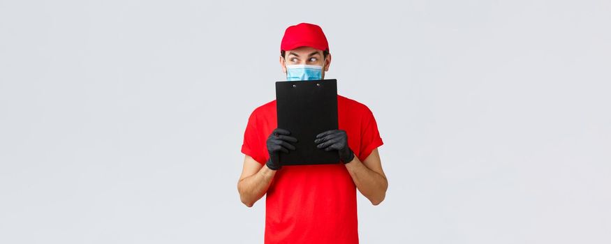 Packages, delivery, covid-19 quarantine, transfer orders. Curious and excited courier in red uniform, gloves and face medical mask, look away scheming, holding clipboard with order form.