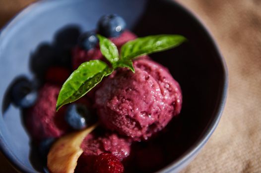 Food still life. Top view. Close-up. Refreshing homemade raw vegan huckleberry ice cream sorbet, melting on a navy ceramic bowl, ornated with lemon basil leaves, blueberries and ripe juicy peaches.