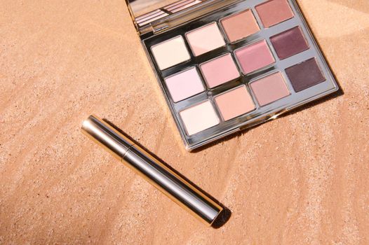 View from above of glamour, luxury eyeshadow palette with gradient from dark brown to shades of beige and mascara on golden sand background with advertising copy space. Summer fashion. Makeup concept.