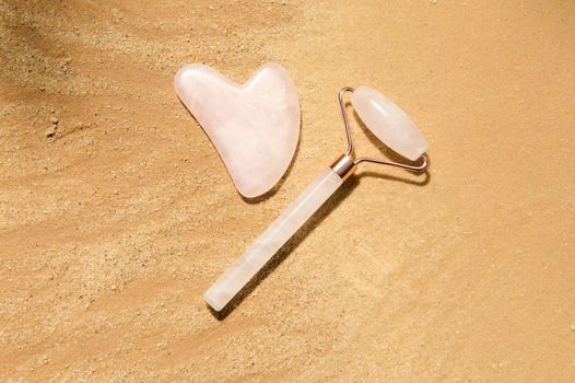 Flat lay. Minimalist still life art of a jade roller of pink stone and gua-sha massager, for home face lifting and lymphatic drainage massage, isolated on a golden sand background with copy ad space