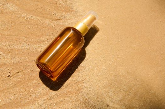 Top view. Flat lay. Minimalist still life of unbranded dark glass bottle with SPF oil or sunscreen body lotion on golden sand beach background. Summer cosmetic concept. Copy space for advertising text