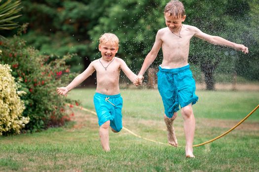 Kids play with water on hot summer day. Children with garden sprinkler having fun. Outdoor fun. Boys run on a field under water drops.