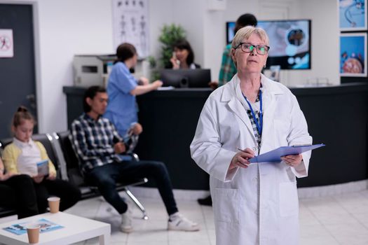 Portrait of older doctor with stethoscope holding clipboard with patient data in hospital reception looking at camera. Confident senior medic in clinic reception lobby waiting for patient appointment.