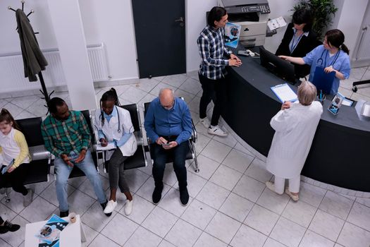 Top view of hospital waiting area with senior doctor admitting asian patient into clinic at front desk. African american doctor filling form for patient appointment in medical healthcare facility.