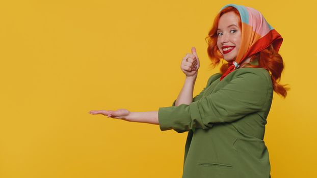 Beautiful redhead woman showing thumbs up, pointing empty place, advertising area for commercial text, copy space for goods promotion. Young ginger girl. Studio shot indoors on yellow wall background