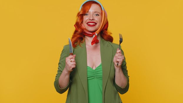 Ready to eat. Hungry redhead young woman in green jacket and dress waiting for serving dinner dishes with cutlery, will appreciate delicious restaurant meal. Ginger girl isolated on yellow background