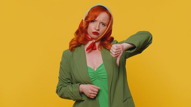 Upset unhappy red hair woman in jacket and kerchief showing thumbs down sign gesture, expressing discontent, disapproval, dissatisfied, dislike. Young ginger girl. Indoors studio on yellow background